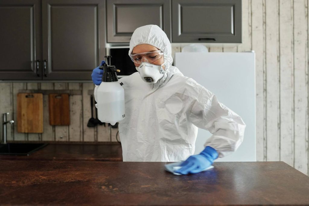 Sanitation worker in PPE spraying and wiping a kitchen counter for disinfection