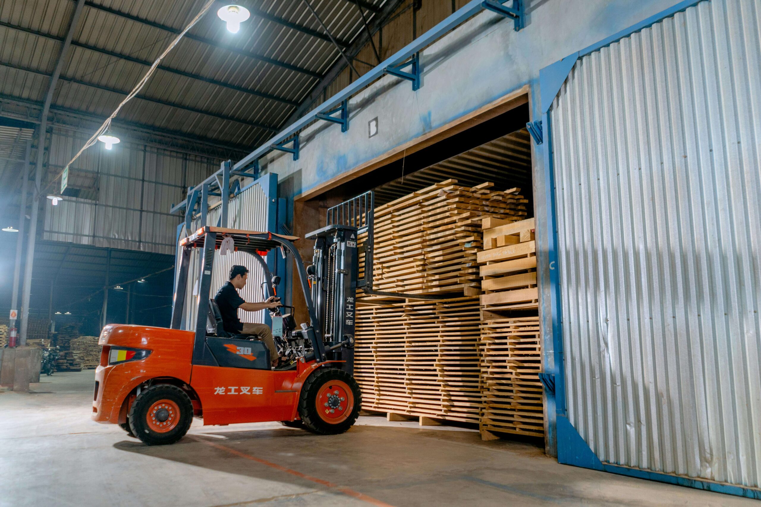 A forklift operator moves stacked lumber planks inside an industrial warehouse facility